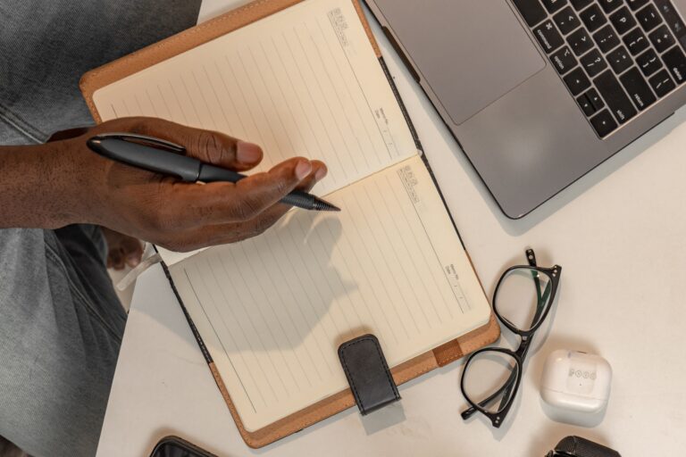 Man writing in a notebook at desk with laptop and eyeglasses. Organized workspace concept.